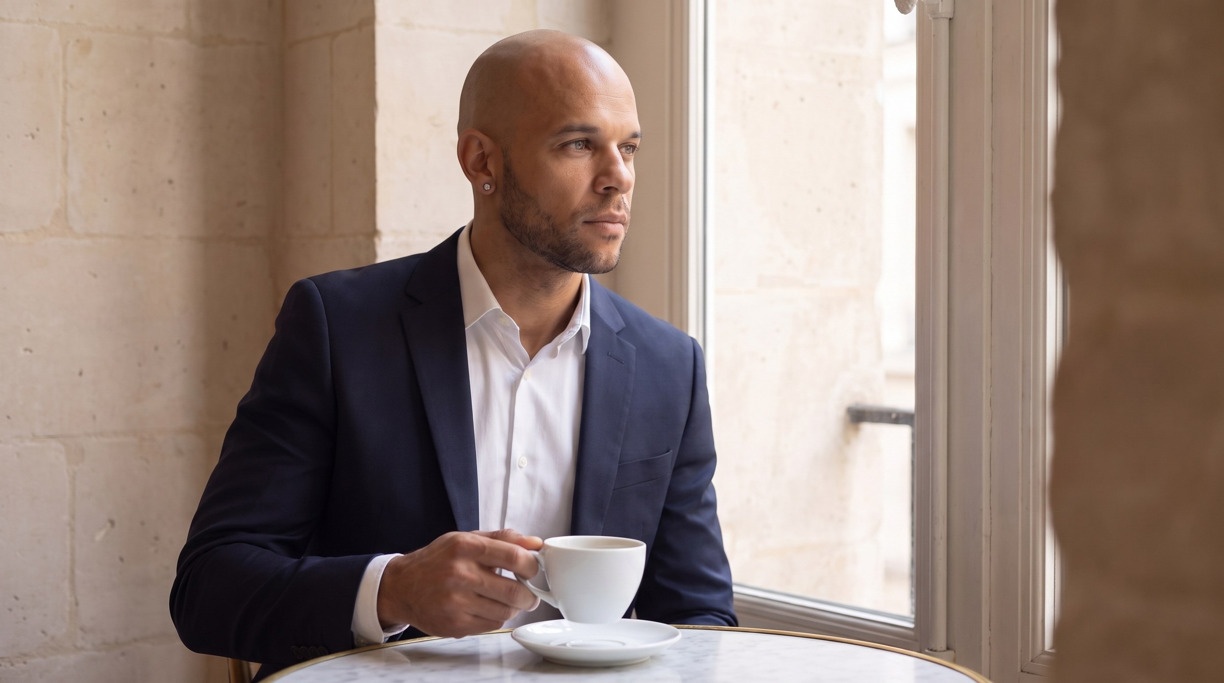 JT Taylor MS, MBA, founder of Notebook of a COO, seated by a window in a navy suit with a cup of coffee
